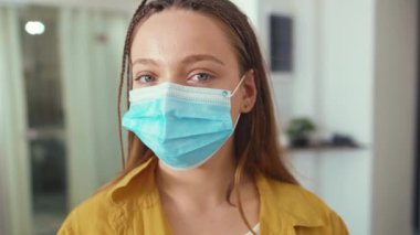 Yüzünde tıbbi maske olan genç bir kadın Coronavirus salgınıyla evinde kamera covid-19 karantina salgınına bakıyor.