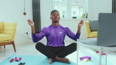 Afrikalı Amerikalı bir adam Lotus 'ta oturmuş bilgisayarının yanında poz veriyor. İnternetteki yerde tek başına meditasyon yapıyor. Yoga sağlık fitness teknolojisi yavaş çekim çalışmasından sonra internetten gözlerini kapatıyor.