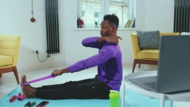 Ciddi Afrikalı Amerikalı bir adam evde dizüstü bilgisayar kullanarak spor salonunda spor yapmak için lastik bant kullanıyor spor kıyafetleri spor kıyafeti spor salonu aktivitesi yavaş çekim