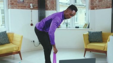 Çekici Afrikalı Amerikalı bir adam online antrenörle egzersiz yapıyor. Evde dizüstü bilgisayar kullanıyor. Spor salonunda spor amaçlı lastik bantlar kullanıyor.