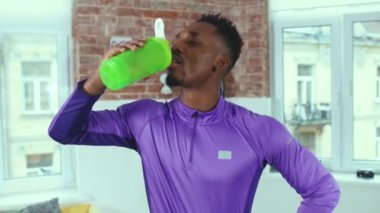 Spor kıyafetli Afrikalı Amerikalı bir adam dinleniyor ve odasında su içiyor. Gülümseyen spor salonu spor salonu, yavaş çekimde kapanıyor.