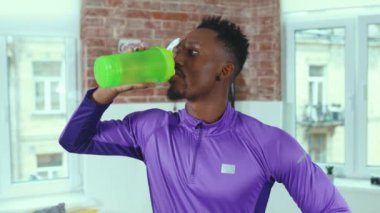 Spor kıyafetli Afro-Amerikan bir adam, dinleniyor ve odasında su içiyor. Gülümseyen spor salonu spor salonu yavaş çekim kapanıyor.
