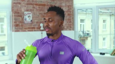 Spor kıyafetli Afro-Amerikan bir adam dinleniyor ve odasında su içiyor. Kamera fitness spor salonuna bakın. Ağır çekim yakın çekim.