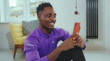 Mutlu Afrikalı genç Amerikalı adam gülümsüyor. Spor eğitimi aldıktan sonra yere oturup rahatlıyor. Cep telefonu akıllı telefon portresi.