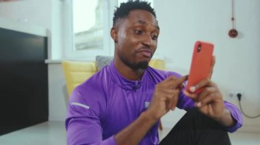 Komik genç Afrikalı Amerikalı adam telefon tebessümleri kullanıyor. Spor eğitimi aldıktan sonra yere oturup rahatlıyor. Cep telefonu akıllı telefon portresi.