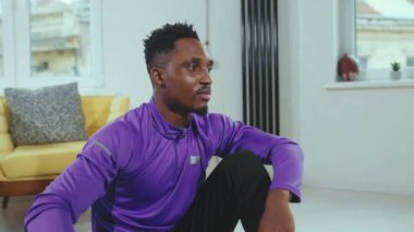 Gülümseyen Afro-Amerikan erkeği, modern apartman dairesinde aldığı eğitimden sonra kameraya bakıyor. Sağlıklı, kararlı, ağır çekimde.