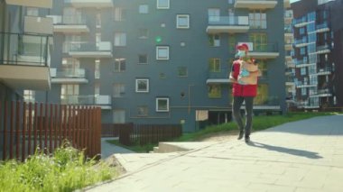 Üniformalı genç kurye tıbbi maske takıyor. Cep telefonu kullanıyor. Karantina sırasında içinde yiyecek dağıtım işçisi olan bir paket arıyor.