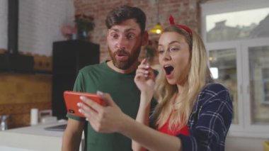 Mutlu çift akıllı telefon kullanıyor, gülümsüyor, ekrandaki telefona bakıyor ve kadın sosyal medya dairesinde mutfakta duruyor, erkek uygulaması yavaş çekim.