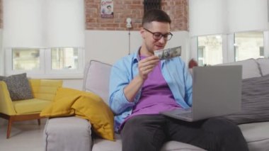Komik genç adam internette para kazanmak için dizüstü bilgisayar kullanıyor. Kanepede el sıkışarak dolar sallıyor. Serbest portre, online piyango yatırımı.