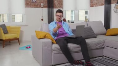Kanepede oturan çekici, serbest çalışan genç bir adam dizüstü bilgisayarını kullanıyor. Evde kalıp bilgisayarını işaret ediyor. Kendini tecrit eden online öğrenci teknolojisini evden yavaş çekimde kullanıyor.