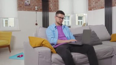 Kanepeye oturup elini sallayan serbest çalışan bir adam. Evde kalıp kendini izole eden online öğrenci teknolojisini yavaş çekimde kullanıyor.