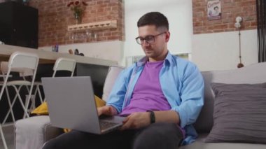 Kanepede oturan genç serbest çalışan dizüstü bilgisayar evde başparmağını gösteriyor. Kendini tecrit eden internet teknolojisi evden yavaş çekimde çalışıyor.