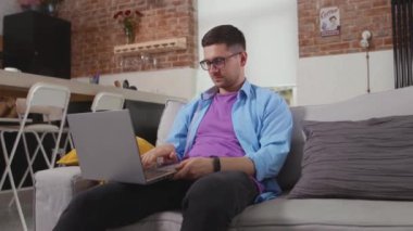 Kanepede oturan serbest çalışan bir adam dizüstü bilgisayarı gösteriyor. Evde çalışan kendini tecrit teknolojisi yavaş çekimde.