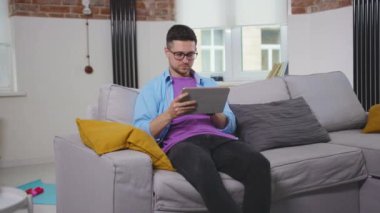 Genç adam evinde tablet bilgisayar kullanıyor. Kanepeye oturmuş gülümsüyor. Günümüz modern karantinasını kapatıyor.