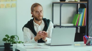 Gözlüklü çekici genç adam dizüstü bilgisayarlı iş yeri karantinası bilgisayar hijyenik ofis çalışanı ağır çekimde çalışmadan önce antiseptikle elleri dezenfekte ediyor.