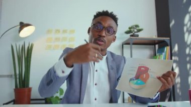 NEW YORK - 5 Nisan 2020: Gözlüklü Afro-Amerikan iş adamını kapatın, istatistikler video blogu çeken veya Webinar sözcüsü olan şirket çalışanlarıyla konuşurken istatistikleri gösteriyor