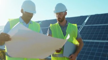 Güneş ışığında güneş çiftliğinde proje planına sahip iki mühendis ayakta durmuş güneşli pillerin kurulumunu tartışıyorlar. Elektrik yavaş çekim.
