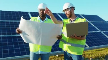 İki genç mühendis güneş çiftliğinde dizüstü bilgisayar ve proje planı kullanıyor. Güneş pillerinin montajını tartışıyorlar. Elektrik yavaş çekim.
