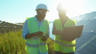 Ağır çekimde iki adam güneş çiftliğinde dizüstü bilgisayar kullanıyor. Özel üniformalı yürüyorlar. Güneşli bataryaların kurulumunu tartışıyorlar.