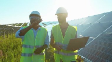 Genç iki adam güneş çiftliğinde dizüstü bilgisayar kullanıyor. Güneşli bataryaların kurulumunu tartışıyorlar. Afrika kökenli Amerikan elektriği yavaş çekim.