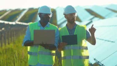 Güneş ışığına yakın adamlar güneş çiftliğinde dizüstü bilgisayar kullanıyorlar. Özel üniformalı yürüyorlar. Güneşli bataryaların kurulumunu tartışıyorlar.
