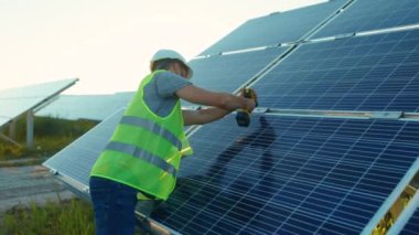 Genç bir işçi güneş panelini güneşli bir günde yeşil bir çatı mühendisi fotovoltaik elektrik bataryasıyla metal bazlı matkapla tamir ediyor.
