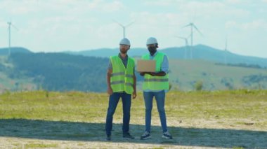 Portre genç mühendisleri tarla ekibinde yel değirmenlerinin yanında dizüstü bilgisayar standı kullanırlar iş birliği hakkında tartışırlar rüzgar enerjisi tarım teknisyenleri yavaş çekimde konuşurlar