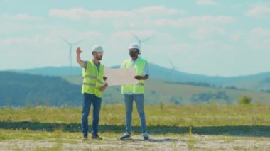 Mühendisler proje planıyla birlikte sahada yel değirmenlerinin yanında durup ekip çalışması yapıyorlar. Rüzgâr enerjili tarım teknisyenleri yavaş çekim konuşuyorlar.