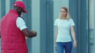 Afrikalı Amerikalı teslimatçı paketçi kadın müşteri. Paket taşıma üniforması için bağlantısız terminalden ödeme yapıyor. Dışarıdaki hızlı sipariş işçisi yavaş çekim kapatıyor.