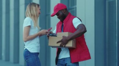 Afrikalı Amerikalı teslimatçı paketçi kadın müşteri. Paket için telefonla ödeme yapıyor. Üniformalı özel sipariş işçisi dışarıda yavaş çekimde kapatıyor.