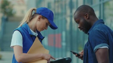 Mavi şapkalı, gülümseyen genç kadın kurye. Afro-Amerikan müşterisi. Paket taşıma üniforması için telefonla ödeme yapıyor.