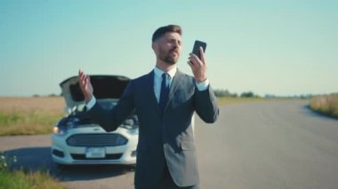 Sinirli iş adamı yalnız başına duruyor. Bozuk arabanın yanında telefonla konuşuyor. Kaput yolunu açıyor.