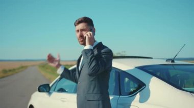 İş elbiseli kızgın adam, yolda akıllı telefonla konuşurken arabanın yanında duruyor ve güneş ışığında iş görüşmesi yapıyor. Yakışıklı telefon işi ağır çekim.