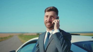 Takım elbiseli genç adam yolda akıllı telefonla konuşurken arabanın yanında duruyor ve güneş ışığında iş görüşmesi yapıyor. Yakışıklı telefon işi ağır çekimde.