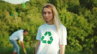 Çekici genç kadın kameraya gülümse beyaz tişört giyip geri dönüşüm işareti takın aile ekolojisi ekolojik toplum eldivenleri yavaş çekim