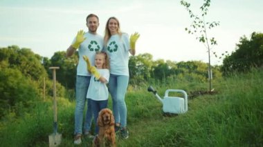 Köpek ekili genç bir aile el sallarken kameraya bakın. Birbirlerine sarılıyorlar. Yeşil bahçeli adam.