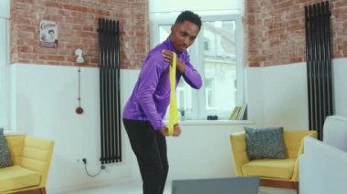 Çekici Afrikalı Amerikalı bir adam online antrenörle egzersiz yapıyor. Evde dizüstü bilgisayar kullanıyor. Spor salonunda spor amaçlı lastik bantlar kullanıyor.