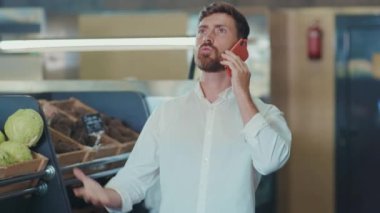 Kızgın genç adam süpermarkette durup telefonda konuşuyor. Sinirli alıcılar, müşteriler, yemek dükkanları, yetişkin pazarları, perakendecilik, yavaş çekim...