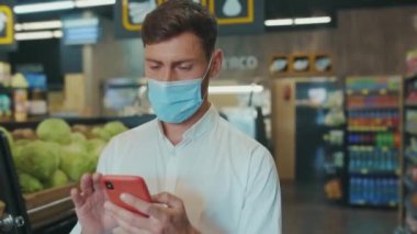 NEW YORK - 19 Nisan 2020: Süpermarkette maskeli genç adam telefondaki alışveriş listesine bakıyor sebze müşterileri için gıda mağazası yetişkin pazarı erkek perakende yavaş çekim
