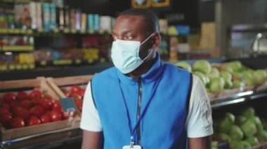 Genç Afro-Amerikan satışları eldiven giyer ve maske takar. Süpermarketteki kameraya bakar. Kara gıda satıcısı.