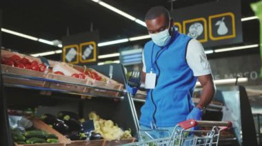 Afro-Amerikan satışlarını kapatın. Eldiven giyin ve maske takın. Maskeli adam süpermarketteki raflara sebze koyar.