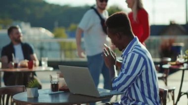 Bir kafede oturan Afrikalı Amerikalı yüksek sesle konuşur. Arka planda dizüstü bilgisayar kullanır. İnsanlar onunla tartışır.