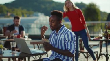 Bir kafede oturmuş hamburger yiyen genç Afro-Amerikalı yüksek sesle arka planda bir dizüstü bilgisayar kullanıyor insanlar onunla tartışıyor ve çok ırklı, yavaş çekimde, yumuşak bir şekilde konuşmak istiyorlar.