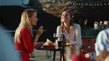 Dinlenen iki kadın telefonda konuşuyor. Kokteyl içiyorlar. Yazlık cafe caddesinde patates kızartması yiyorlar.