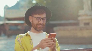 Yavaş çekim genç adam şapka takıyor ve çiçekli gözlük takıyor açık havada erkek arkadaşlarını bekliyor telefonda gülümsüyor eşcinsel arkadaşlık ilişkisi yaşam tarzı konsepti homoseksüel Igbt