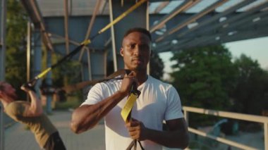 Ciddi Afrikalı Amerikalı adam trx fitness halatıyla dolaştıktan sonra kameraya bakıyor. Stadyumdaki kas sporları spor kıyafetleri spor salonunda vücut egzersizleri. Yavaş çekim yakın çekim.
