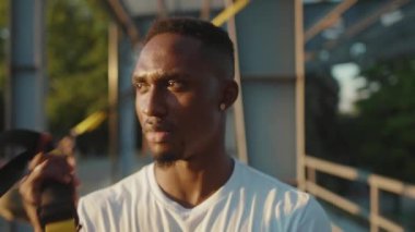 Genç Afrikalı Amerikalı adam trx fitness halatıyla hareket ettikten sonra gülümseyen kameraya bakıyor. Stadyumdaki spor kıyafetleri spor salonunda vücut egzersizleri yapıyor. Yavaş çekimde spor portresi kapanıyor.