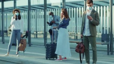 Otobüs durağında, tıbbi maskeli, nakil için bekleyen genç insanlar tren istasyonu gezginleri, gezginler, Coronavirus sosyal mesafe salgınında telefon kullanan kadın ve erkekleri kullanıyor.