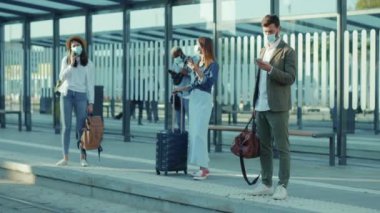 Güneş ışığında, sağlık maskeli, otobüs durağında bekleyen genç insanlar tren istasyonundaki kadın turistleri, Coronavirus sosyal mesafe salgınını yavaş çekimde kullanıyorlar.