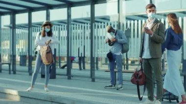 Melez ırkları kapatın. Sağlık maskeli gençler otobüs durağında nakliye için bekliyorlar. Telefoncular, kadın turistler, tren istasyonu gezginleri, Coronavirüs sosyal mesafe yavaş çekim salgını.